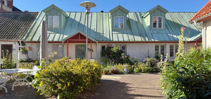 Storgatan 19H, 1 1/2-planshus med vit putsfasad och koppargrönt plåttak. I förgrunden buskar och plattlagd gång.