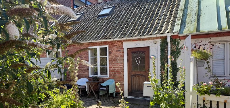 Litet hus i rött tegel med trädörr och vitt sidohängt fönster, med egen uteplats.