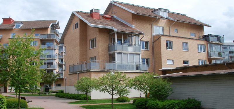 Grönskande träd i förgrunden och bakom ett fyravåningshus i gult tegel med stålfärgade balkongfronter.