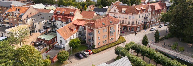 Flygvy över Storgatan 7 sett från sydöst. Trevåningshus plus vindsvåning i orangeroa puts med krämvita fasaddetaljer och brunt tegeltak, samt sidobyggnad i vit puts med beige fasaddetaljer. Vita spröjsade fönster. I förgrunden syns Vita räck och dess grönskande träd, samt den lilla trädgården tillhörande fastigheten med två gräsytor, häckar och träd.