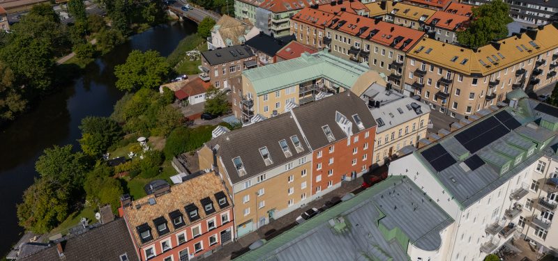 Flygvy från söder, Västra Kyrkogatan 5-7. Två sammanbyggda huskroppar, en i gult tegel och en i brunt. I bakgrunden syns åns naturområde samt grannfastigheter i varierande arkitektur.