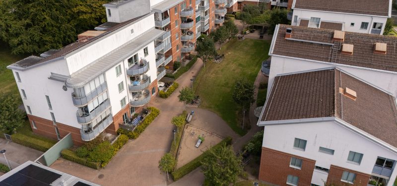 Innergård med plattlagda gångar, gräsyta och lummiga träd samt sandlåda med liten rutschkana, mellan sidobyggnaderna och gårdshusen.