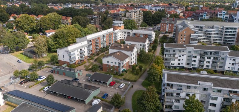 Flygvy från väst där fastighetens runda torn i vinkeln visas i högra hörnet och de två sidobyggnaderna samt gårdshusen syns. Tornet är i brunt tegel och sidodelarna samt gårdshusen är i tegel och vit puts. Bilden visar grannfastigheterna med mellanliggande cykelväg och grönskande innergårdar.