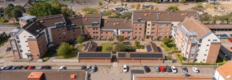 Flygvy över Sockerbruksgatan 12A-F sett från öster. U-formad fastighet med höjder som varierar mellan 5 och 2 våningar. Två L-formade envåningshus beklädda med solceller på tak och grönskande innergård med träd, pergola och sandlåda. Fastigeht är byggd i brunt tegel, med vita och gula detaljer i puts.