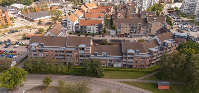 Flygvy över Sockerbruksgatan 12A-F sett från väster. Fastighet i brunt tegel med höjder som varierar mellan 5 och 2 våningar. Indragna och inglasade balkonger mot Industrigatan och grönskande träd och buskar.