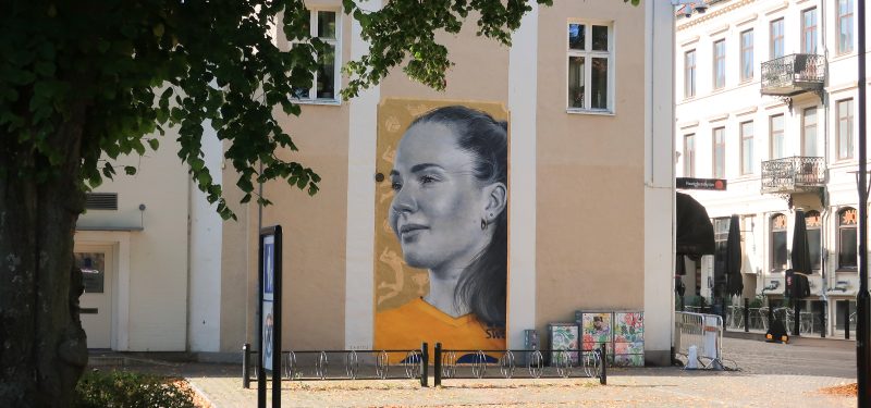 Muralmålning på rosa putsfasad, föreställande volleybollspelaren Isabelle Haak. I förgrunden Stadsparkens träd.