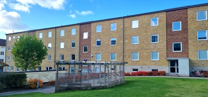 Fasad mot innergård, Bruksgatan 27. Tegelfasad i två olika bruna nyanser, i förgrunden gräsyta. pergola samt grönskande träd.