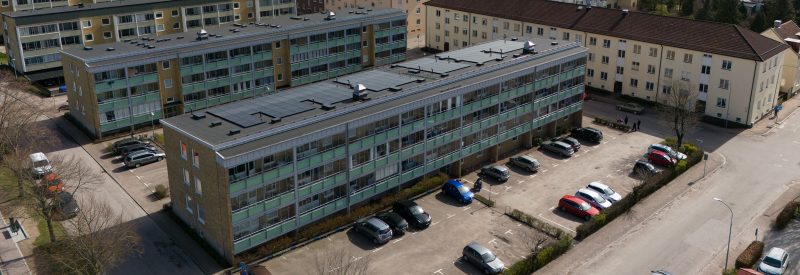 Flygvy över kvarteret Bruksgatan 27-29. Visar två huskroppar i tre våningar med tegelfasad och svart papptak. Huskroppen närmast kameran är försedd med solceller och har en parkering i förgrunden.