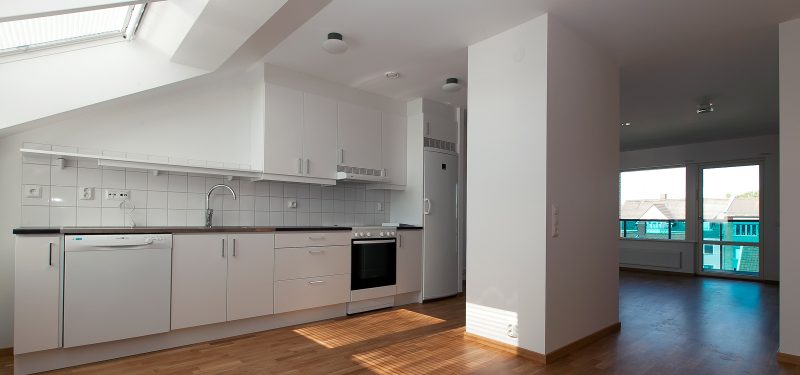 Vit köksinredning med snedtak till vänster med ljusinsläpp från takfönster. Öppen planlösning mot matplats och vardagsrum. Murstock mitt i rummet. Parkettgolv i ek och vita väggar.