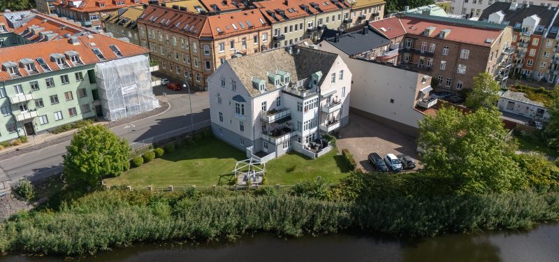 Flygvy sett från ån, äldre hus i tre våningar med balkonger mot ån. Rosa fasad med vita detaljer. Gräsyta, uteplats mot ån.