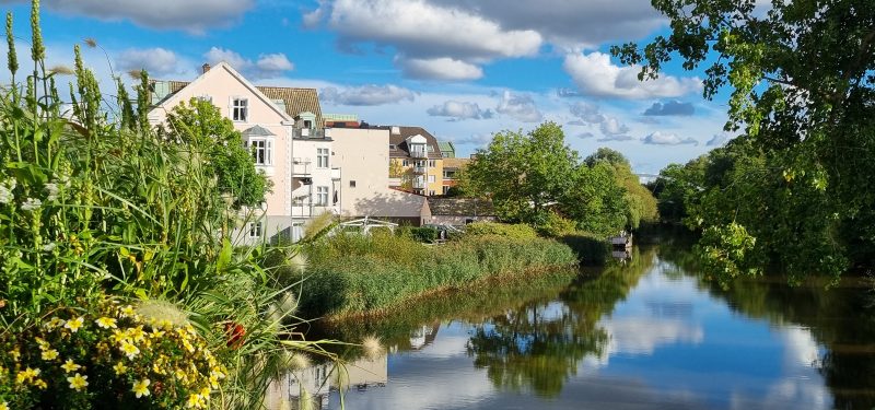 Järnvägsgatan 33 sett från Järnvägsbron. Rönneå och grönskande omgivningar i förgrunden, i bakre del syns det rosa putshuset.