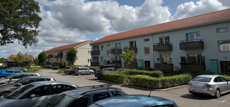 Två tvåvåningsfastigheter med träpanel i bakgrunden med parkeringsplats, uteplats och grönska i förgrunden. Visa Landshövdingevägen 13 och Doktorsgatan 6.