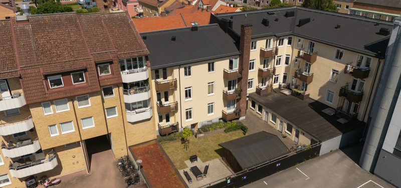 Kv. Strutsen 7 sett från ovan sydväst, visar Östergatan 59/Vaktgatan 6-8 på hörnet mellan två grannfastigheter. Innergården har en gräsyta, träd, perennplanteringar samt uteplats med grill och cykelskjul. Bostadshuset har gul puts samt svart plåttak, med utanpåliggande balkonger med bruna plåtfronter.