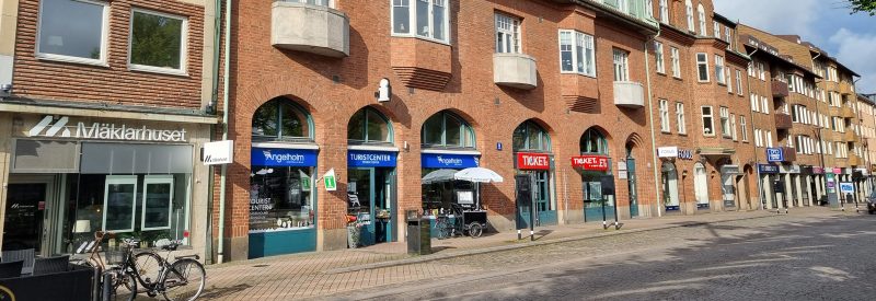 Gatuvy över Järnvägsgatan 5-7 i Ängelholm. Byggnaden har en röd tegelfasad med bågformade fönster och ett grönt plåttak under en blå himmel.