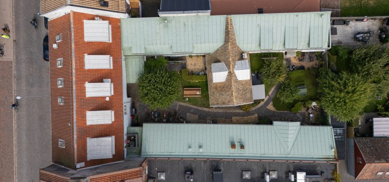 Flygvy över Storgatan 64, sett rakt uppifrån. Visar gatuhusets bruna tegeltak med takkupor mot gården, och gårdshusens koppargröna plåttak i två flyglar. Mitt på övre gårdshuset ligger en byggdel i vinkel med ljust tegeltak och kupor åt båda hållen. Innergård med gräsyta, lummiga träd samt stenlagd gång.
