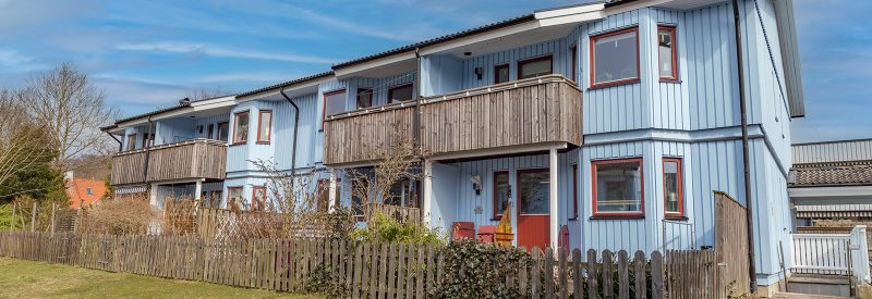 Byggnad i två plan med ljust blå träpanel och röda fönster och dörrar. Balkonger med fronter i tryckimpregnerat trä på våning två och inhägnade uteplatser på bottenvåning.