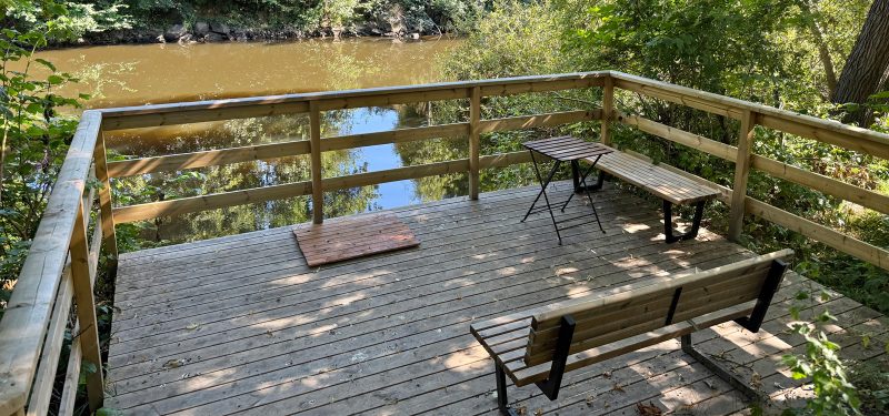 Brygga i trall som omgärdas av steket i tryckt trä, med två sittplatser, i bakgrunden Rönneå och omgivande grönska.