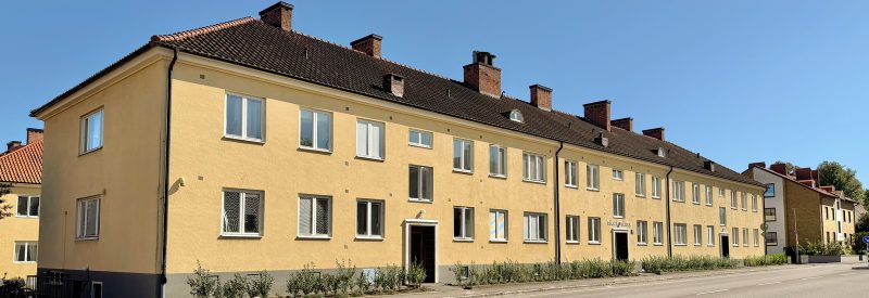 Flerbostadshus i två våningar plus vind, med gul putsfasad, vita sidohängda fönster och brunt tegeltak. Längs hela fasaden växer en häck.