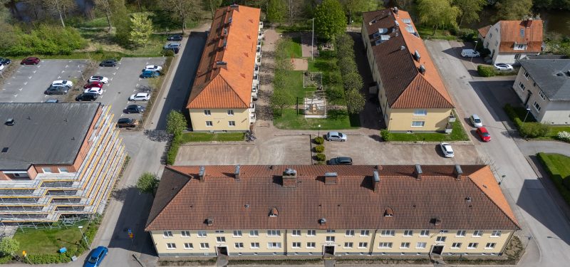 Flygvy över kvarteret Ugglan. Sett från Kristian II:s väg, tre likvärdiga gula putshus i två plan ligger som en u-formation kring en parkering med grusbeläggning och innergård med gräsyta, träd, uteplats under pergola samt sandlåda med gunga.