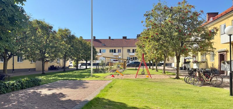 Innergård med boulebana i nedre vänster hörn, stor gräsyta till höger och längs med finns rönnar. Sandlåda med gunga, flaggstång. I bakgrunden syns pergola med uteplats och i horisonten det gula putsade huset mot gatan.