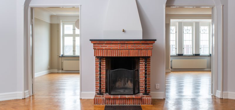 Interiör från en lägenhet, öppen eldstad i tegel, med smidesdetaljer. I bakgrunden syns dörröppningarna in mot vardagsrum och sovrum, med ljusinsläpp från spröjsade fönster. Parkettgolv i fiskbensmönster.