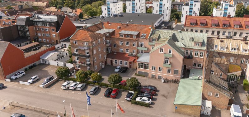 Flygvy över baksida och parkering med grönskande buskar och träd sett från väst. Bostadsfastighet i totalt 4 våningar, där en del har brun tegelfasad och den andra är rosa puts med vita detaljer och koppargrönt plåttak. Ett antal balkonger med staket eller grön plåtfront.