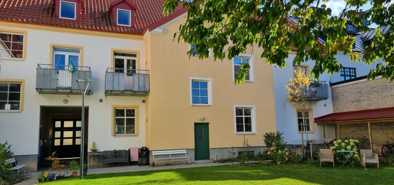 Fastighet i tre våningar med vitputsad fasad med gult parti, balkonger mot grönskande innergård med stort träd i förgrunden.