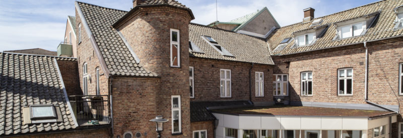 En stor, äldre byggnad i brunt tegel med flera våningar och branta tegeltak, inklusive ett litet runt torn för trapphuset. Fönstren har vita ramar och himlen är ljusblå med några moln.