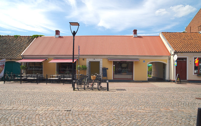 En gul envåningsbyggnad med rött plåttak, identifierad som "Ostbutiken", har en uteplats med bord och stolar på ett kullerstensgata. Framför byggnaden står några cyklar parkerade. Blå himmel med vita moln syns ovanför.