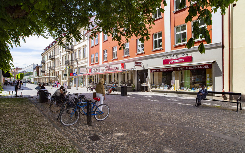En solig gatuscen på Storgatan i Ängelholm med flera affärsfasader, människor som promenerar och sitter på bänkar samt parkerade cyklar i förgrunden.
