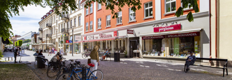 En solig gatuscen på Storgatan i Ängelholm med flera affärsfasader, människor som promenerar och sitter på bänkar samt parkerade cyklar i förgrunden.