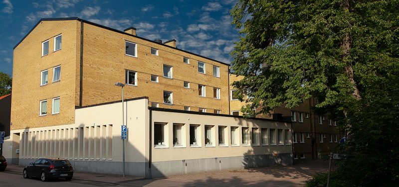 Fastighet i fyra våningar, kläd i gult tegel. Bottenplan har mindre utbyggnad för lokal, denna är putsad gräddvit. I förgrunden skymtar gröna träd mot ån.