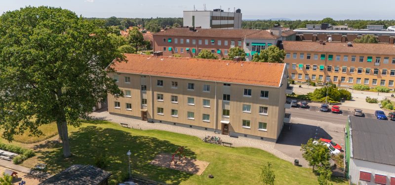 Flygvy över innergård från öst. Stor gräsyta med sandlåda och gunga samt träd, plattlagd gång till entréer och cykelställ. I bakgrunden skymtar Ängelholms sjukhus.