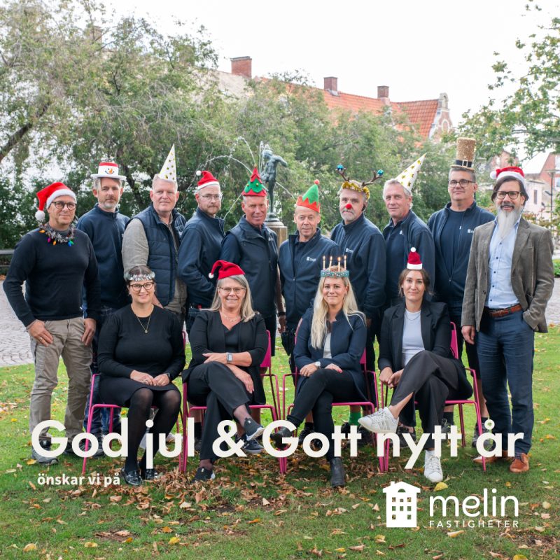 En grupp om tolv personer, åtta män stående i bakgrunden och fyra kvinnor sittande i förgrunden, poserar för ett gruppfoto utomhus i en park. Alla har fått en julig hatt påklistrad, en man har även fått ett tomteskägg. Text: god jul och gott nytt år önskar Melin Fastigheter.